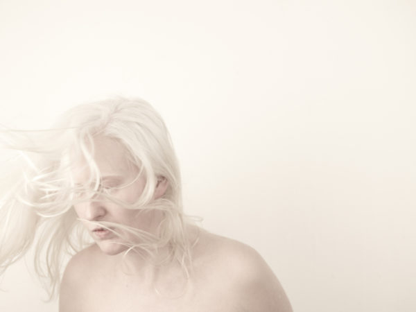 An artist with very white hair and pale skin looks away, pictured against a stark white background