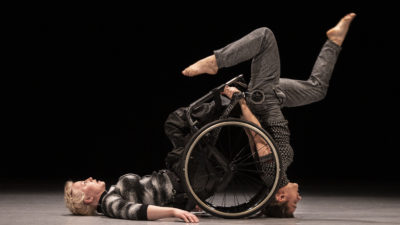 Two female performers, one using a wheelchair lies with her back on the floor, and the other stands on her head with her legs in the air above the chair.