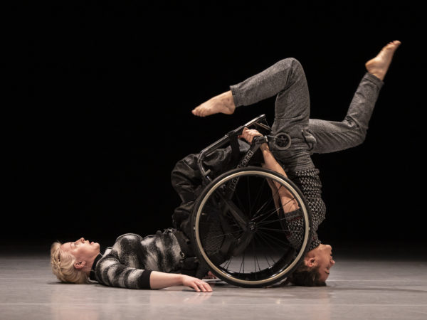 Two female performers, one with blonde hair uses a wheelchair and lies with her back on the floor, the other stands upside down with her legs cycling in the air