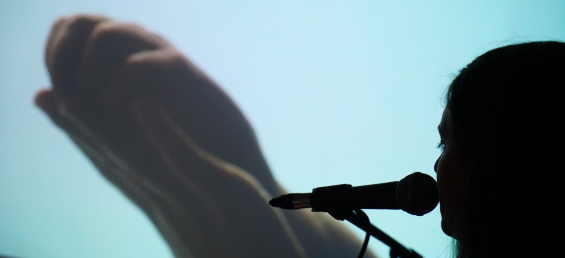 Projection of two hands together with blue background, in foreground is head of female performer facing the projection with microphone.