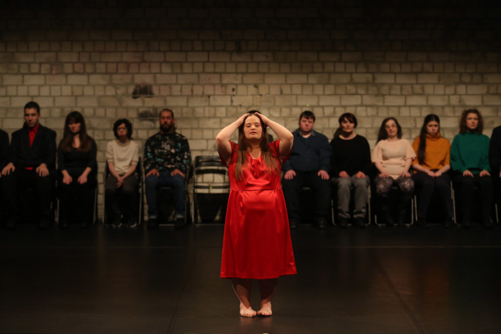 Female performer in red dress, group of other performers sit behind her