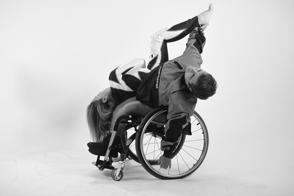 Two female dancers duet, one using a wheelchair and reaching with their hand straight up to catch the foot of the other dancer whose in an upside-down position