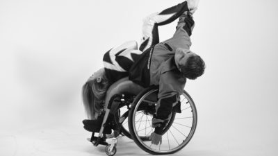 Two female dancers duet, one using a wheelchair and reaching with their hand straight up to catch the foot of the other dancer whose in an upside-down position
