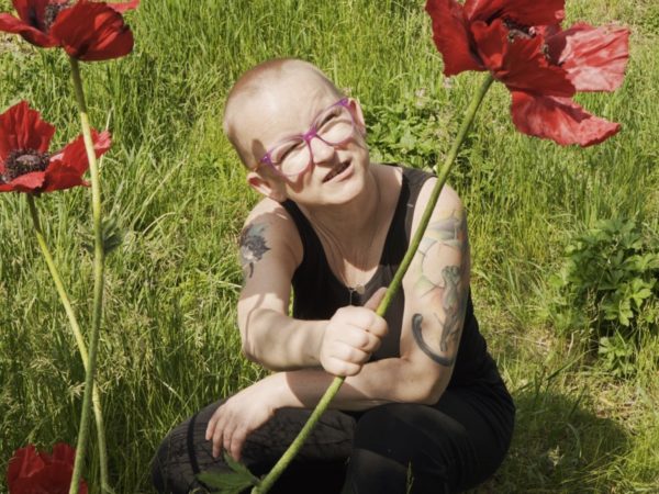 White woman with shaved head and glasses crouching in a field holding a red flower