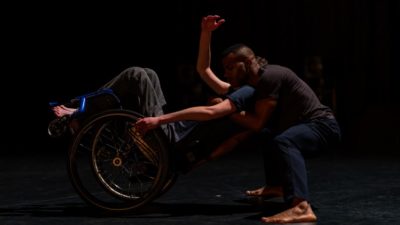 One dancer leans back in a blue wheelchair as another stands behind him to embrace his shoulders.