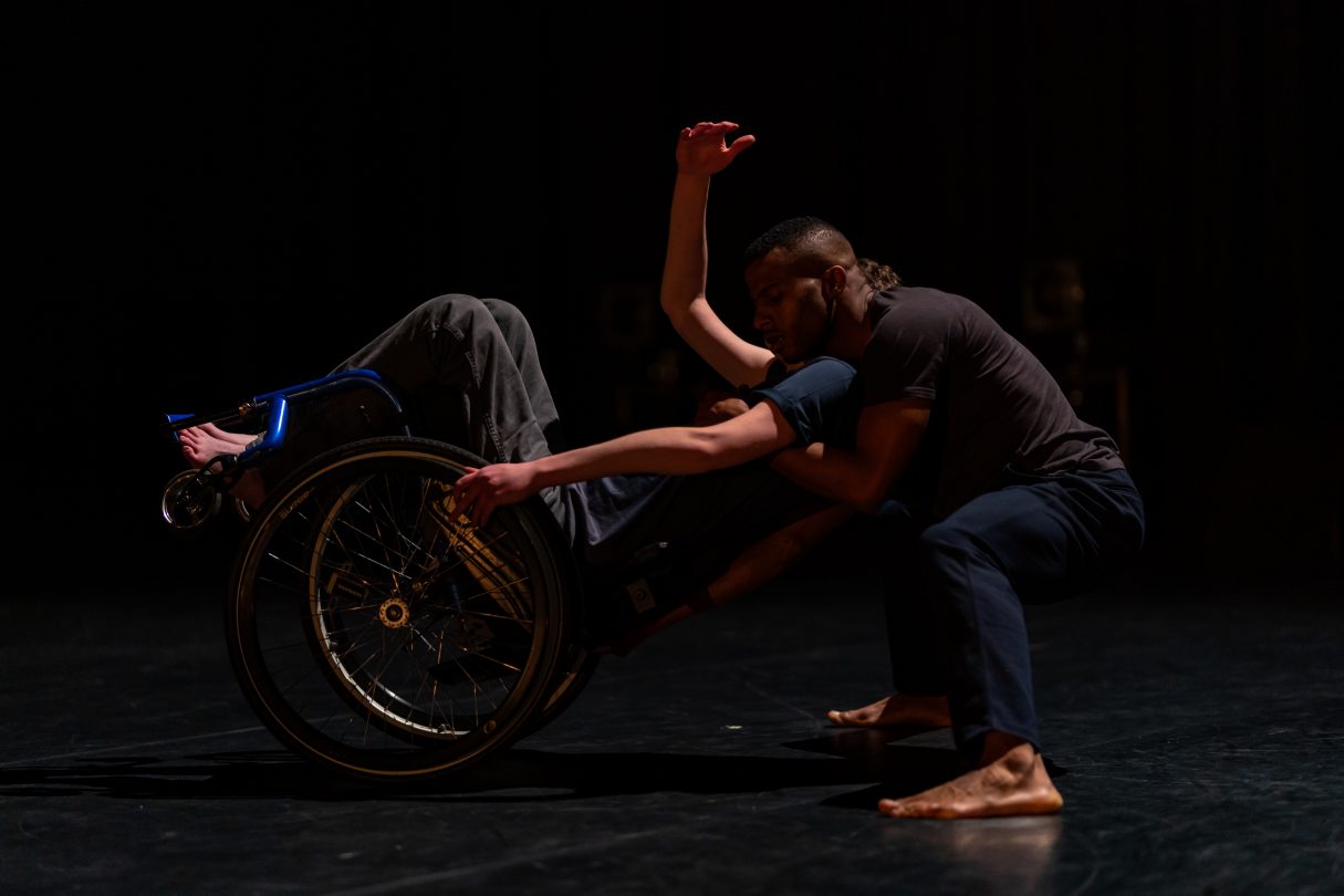 Two male dancers, one leans back in a blue wheelchair, the others crouches low to the floor embracing his shoulders.