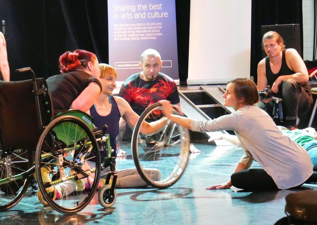 Tatiana sits on the floor having removed a wheel from a wheelchair and spins it with a female Deaf dancer