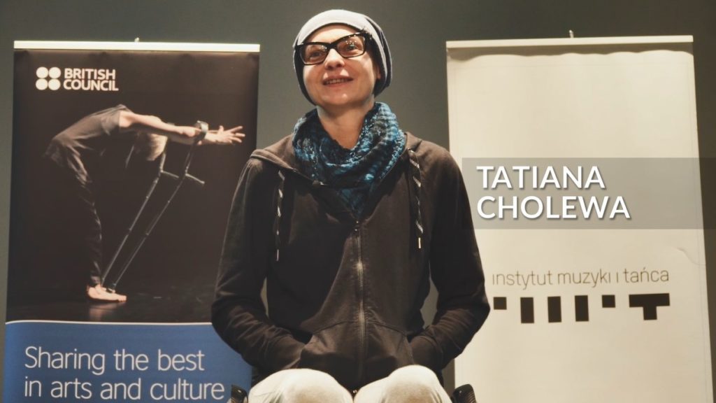 A dancer in a wheelchair smiles in an interview. She is wearing a beanie hat, black rimmed glasses, a black hoodie and a blue patterned scarf.