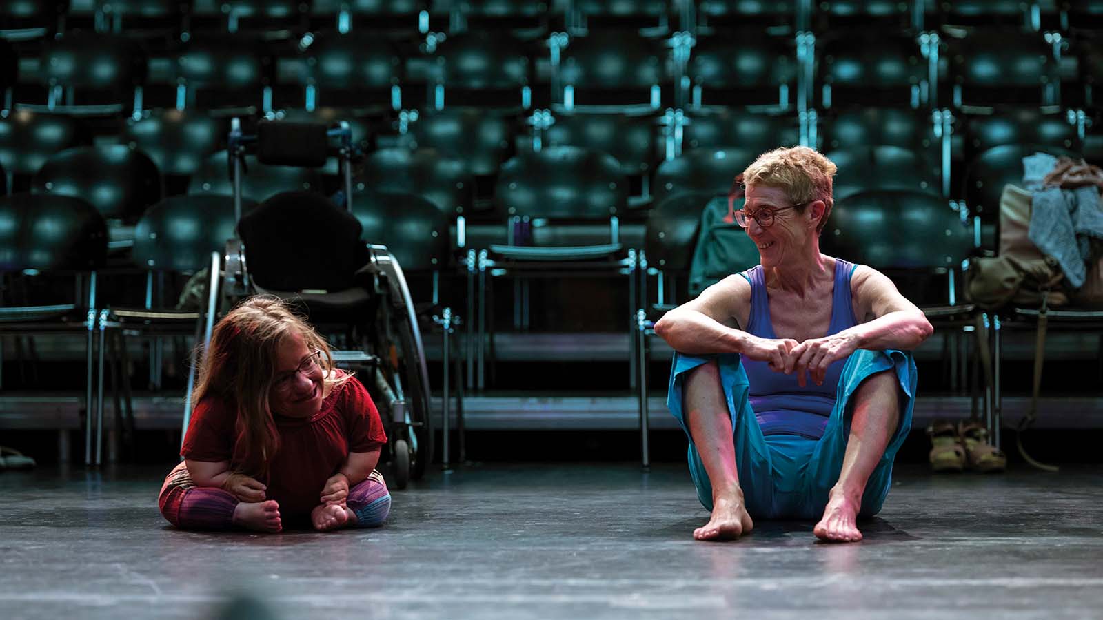 Two female perfromers sit on the floor, one has dwarfism or a similar impairment