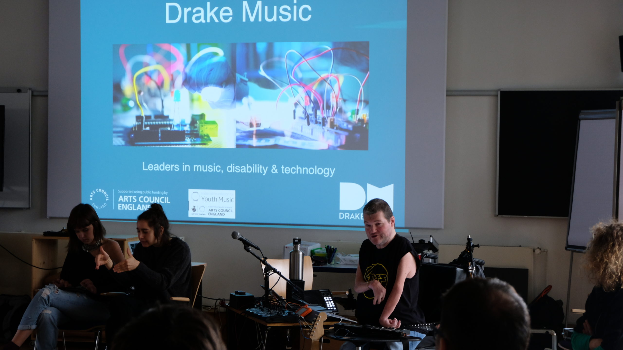 Wheelchair user presenting to a crowd whilst sat in front of music technology equipment with a projection behind him