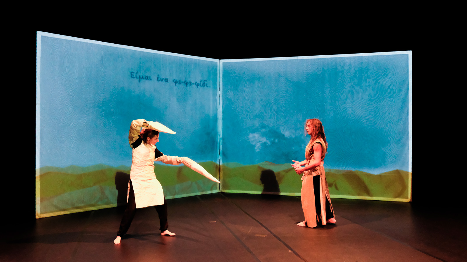 Two performers, on with flippers on their hands, the other in a ripped garment stand against a projected backdrop