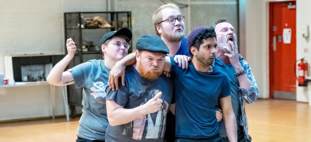 A cast of male actors in rehearsals holding each other in a huddle