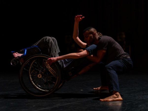A black dancer supports a white dancer who is balancing as they lean backwards on their wheelchair