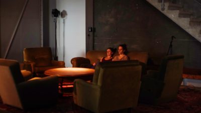 Two female performers sit on a sofa as they create a site-specific work