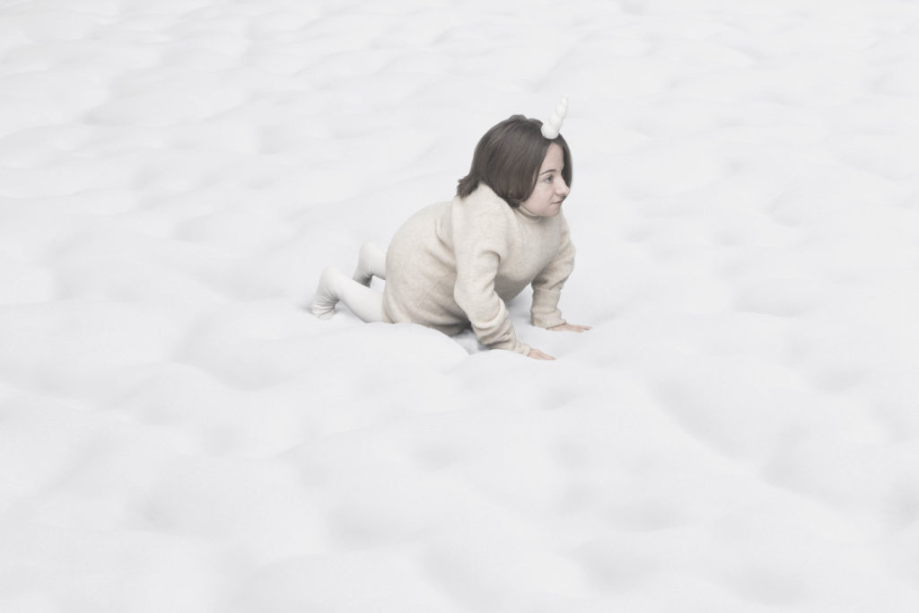 A small female performer all in white with a unicorn's horn crawls across a fluffy cloud landscape.