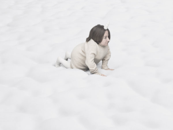 A small female performer crawls across a white blanket of seemingly endless clouds, a unicorn horn on her head.