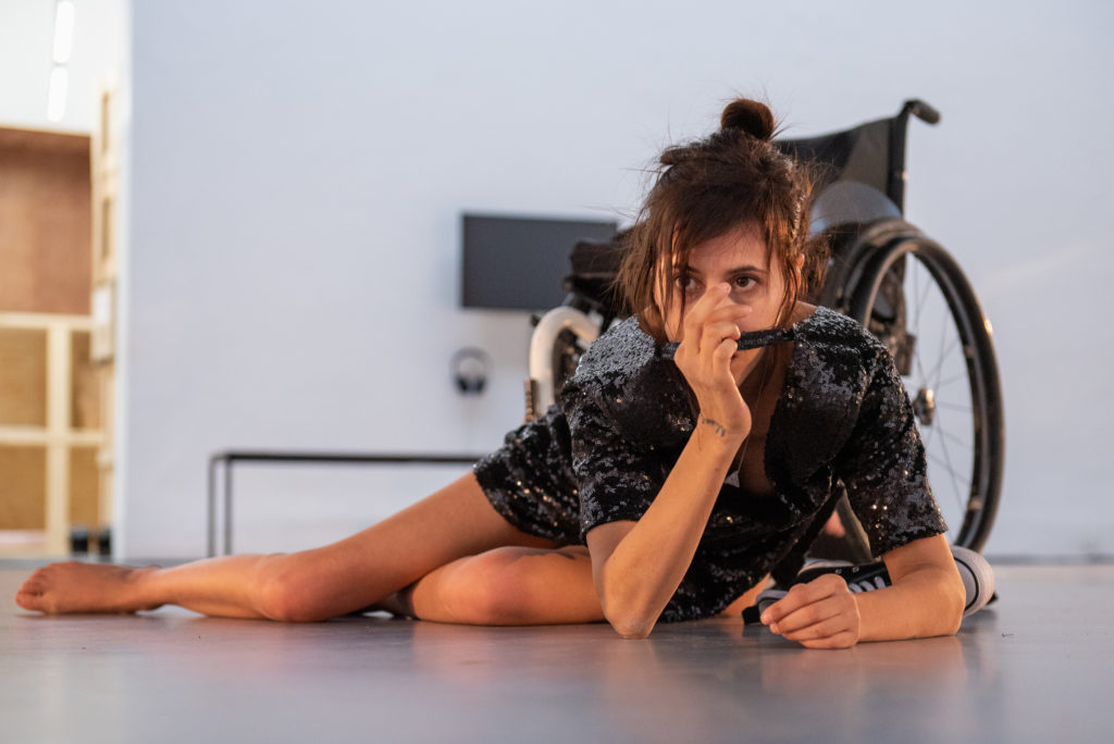 A female performer lays on the floor in front of her wheelchair, wearing a black sequined dress
