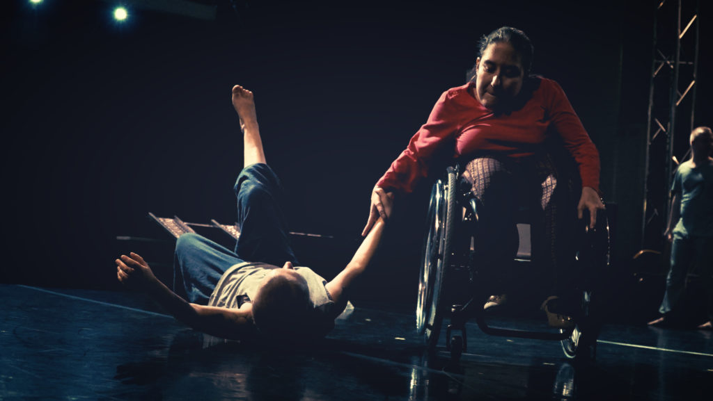 Two performers on a theatre stage, one woman in a wheelchair holds another woman's hand as she lies back on the floor with one leg in the air.