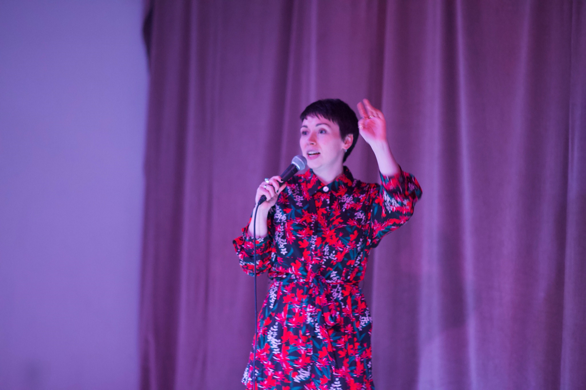 White woman with short dark hair doing stand up comedy, wearing a patterned red and blue outfit