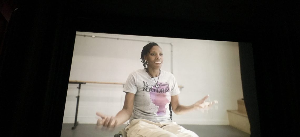 Image of cinema screen shows a female wheelchair dancer mid-interview