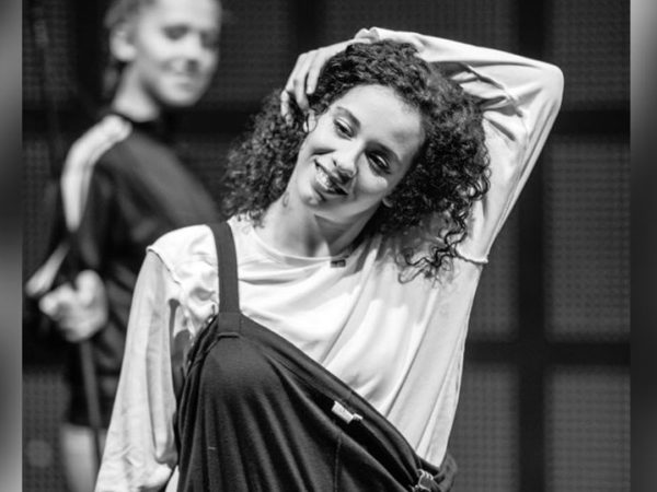 Black and white photo of a female dancer with dark curly hair. She reaches her arm up and over her head, smiling.