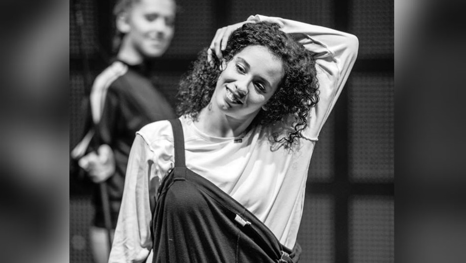 Black and white photo of a female dancer with dark curly hair. She reaches her arm up and over her head, smiling.