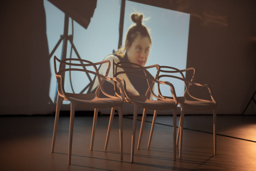 Three chairs at the front of a stage sit before a video projection showing a learning disabled female performer