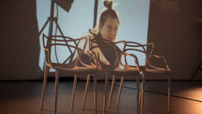 Three chairs at the front of a stage sit before a video projection showing a learning disabled female performer.