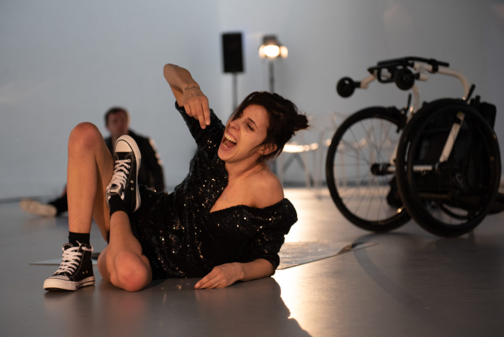 A brunette woman screams on the floor, her wheelchair beside her. She wears Converse trainers and a sparkly black dress.