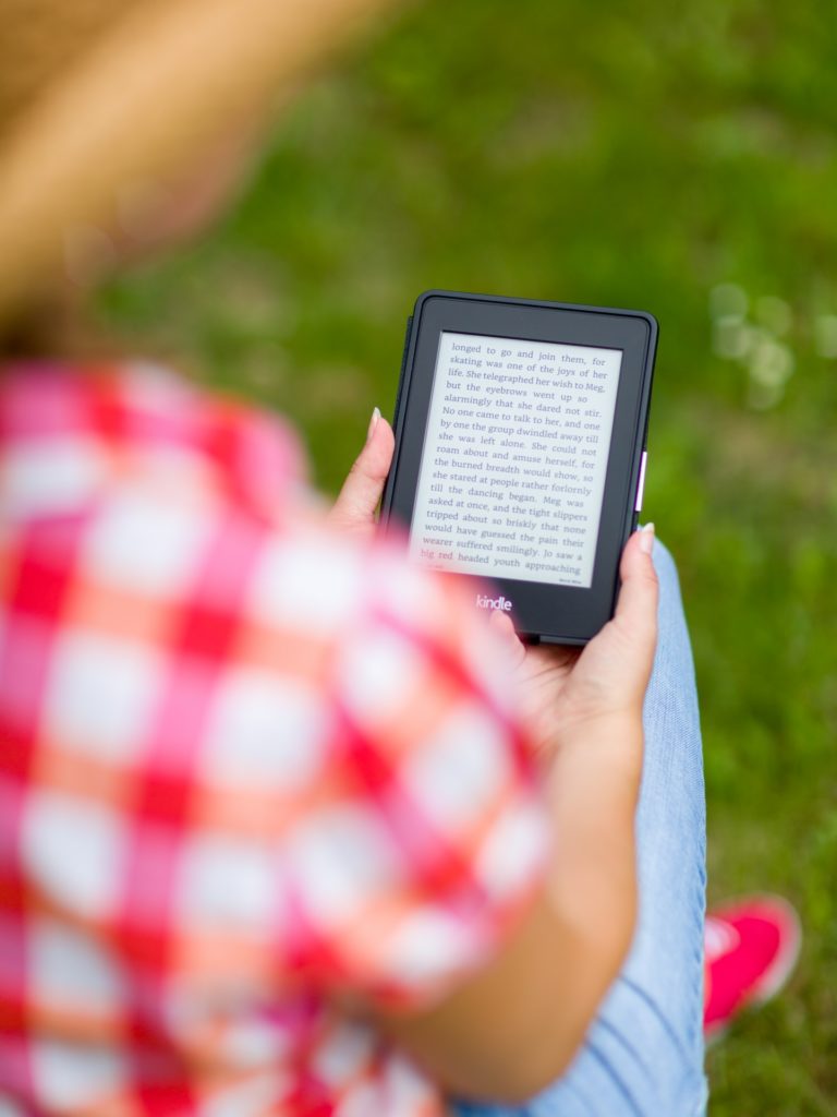 Photo of a person sitting outside holding an e-book reader. The person is out of focus and facing away from the camera, wearing a plaid shirt. The Kindle they hold displays an e-book, and is in sharp focus.