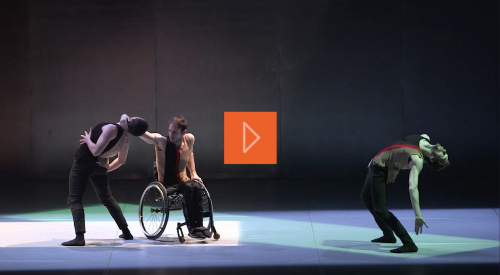 Three male dancer in the same costume, one is a wheelchair user. One is leaning backward almost touching the floor.