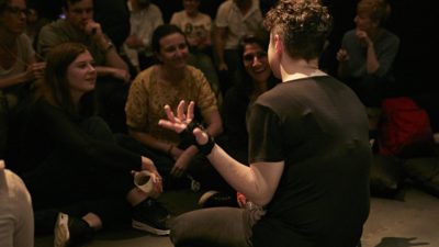 Photograph of Jess Thom, a white woman with curly brown hair sitting on the floor, shot from behind, she is talking to a large audience gathered very close to her