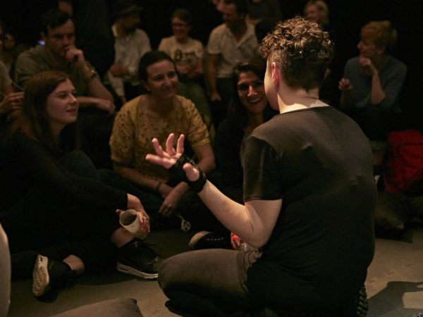 Photograph of Jess Thom, a white woman with curly brown hair sitting on the floor, shot from behind, she is talking to a large audience gathered very close to her