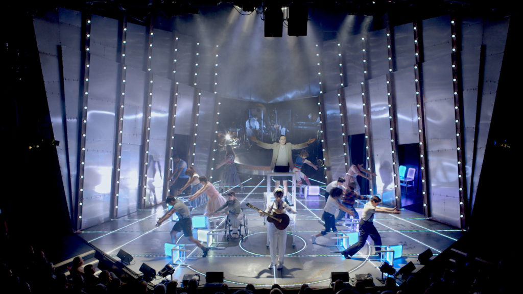 A large choreographed scene of performers on a brightly lit stage 