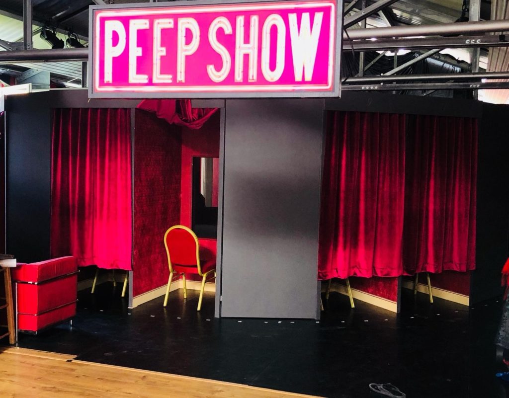 Huge neon pink writing displays the words  'Peep Show'. Underneath sits a red chair facing a hole in the wall and red velvet curtains either side.