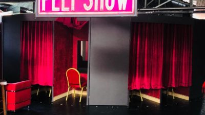 Huge neon pink writing displays the words 'Peep Show'. Underneath sits a red chair facing a hole in the wall and red velvet curtains either side.