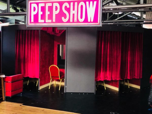 Huge neon pink writing displays the words 'Peep Show'. Underneath sits a red chair facing a hole in the wall and red velvet curtains either side.