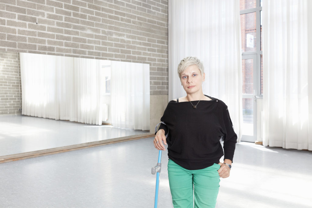 A woman with short cropped light blonde hair stands in a dance studio with a crutch. She is wearing a black top and mint green trousers.