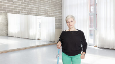 A woman with short cropped light blonde hair stands in a dance studio with a crutch. She is wearing a black top and mint green trousers.