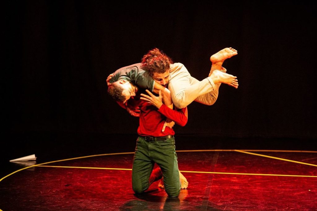 One performer in a red jumper and jeans holds another performer cradled around his shoulders in an embrace.