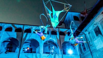 Blue light illuminates an aerial performance suspending performers high in the air in a historic courtyard.