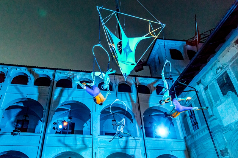 Blue light illuminates an aerial performance suspending performers high in the air in a historic courtyard.