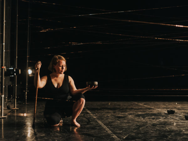 Woman with red hair and a walking stick is kneeling on a black floor in a dark theatre space holding a bowl