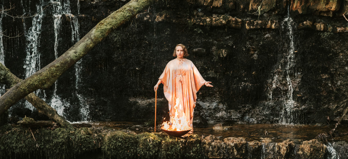 A woman in pink robes stands on the ledge of a small waterfall with a bowl of fire at her feet