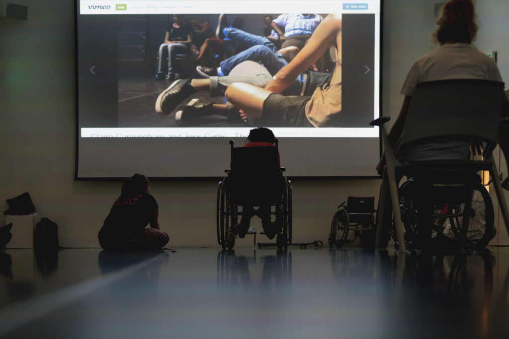 Two wheelchair users and another participants sit viewing a large screen video of a dance performance.