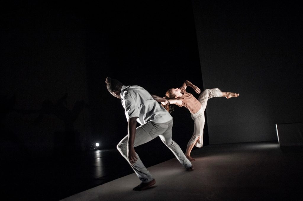 A male and female performer, illuminated in a dark room. The female seems to pull away, her left leg raised high in the air, the male performer counterbalancing her pose.