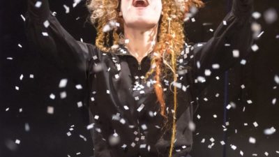 Female performer looks joyously to the sky as snowflakes fall around her She is wearing a big blonde wig and a black dress