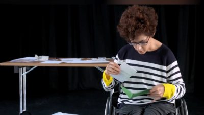 A female performer in a wheelchair sifts through an abundance of letters