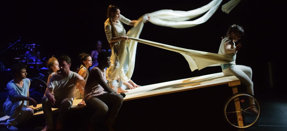 On the top edge of a sloping wheeled table, a performer is tied by a long white cloth and pulled back by another performer who’s grabbing the cloth like a horse-driver.