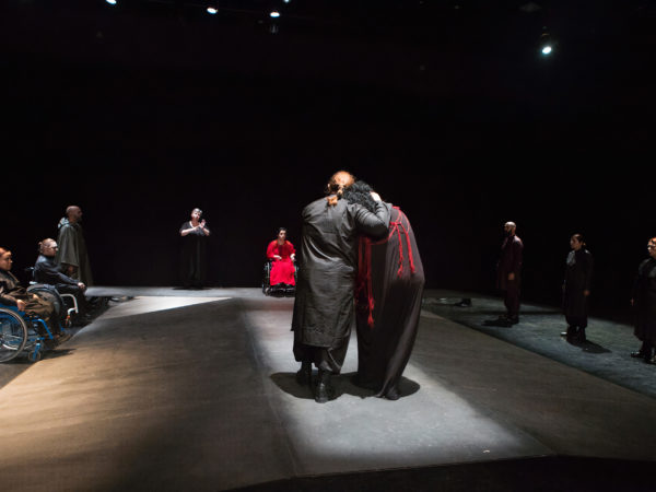 Performers dressed in black form a corridor as two women, one resting their head on the other's shoulder walk through the centre